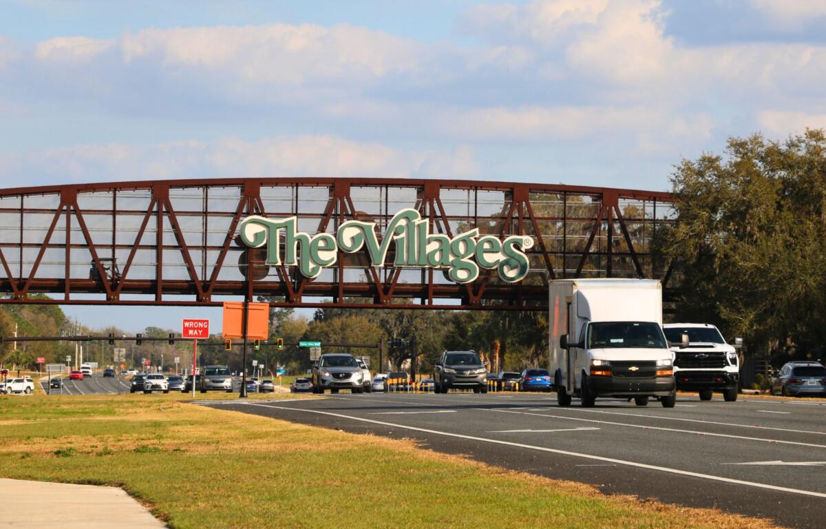 A brown, metal bridge for golf cart passing features large, green letters with white borders reading, "The Villages." A busy road passes underneath the bridge.