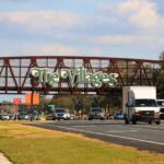 A brown, metal bridge for golf cart passing features large, green letters with white borders reading, "The Villages." A busy road passes underneath the bridge.