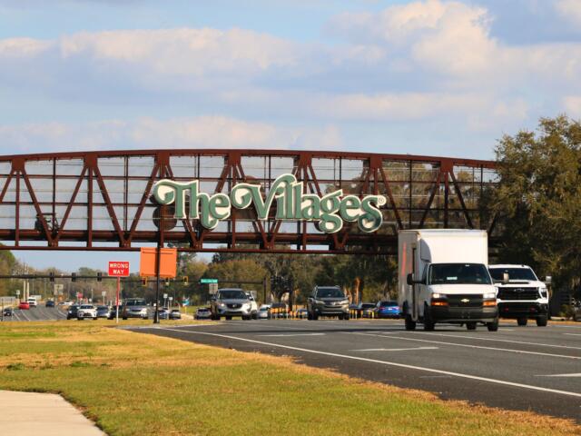 A brown, metal bridge for golf cart passing features large, green letters with white borders reading, "The Villages." A busy road passes underneath the bridge.