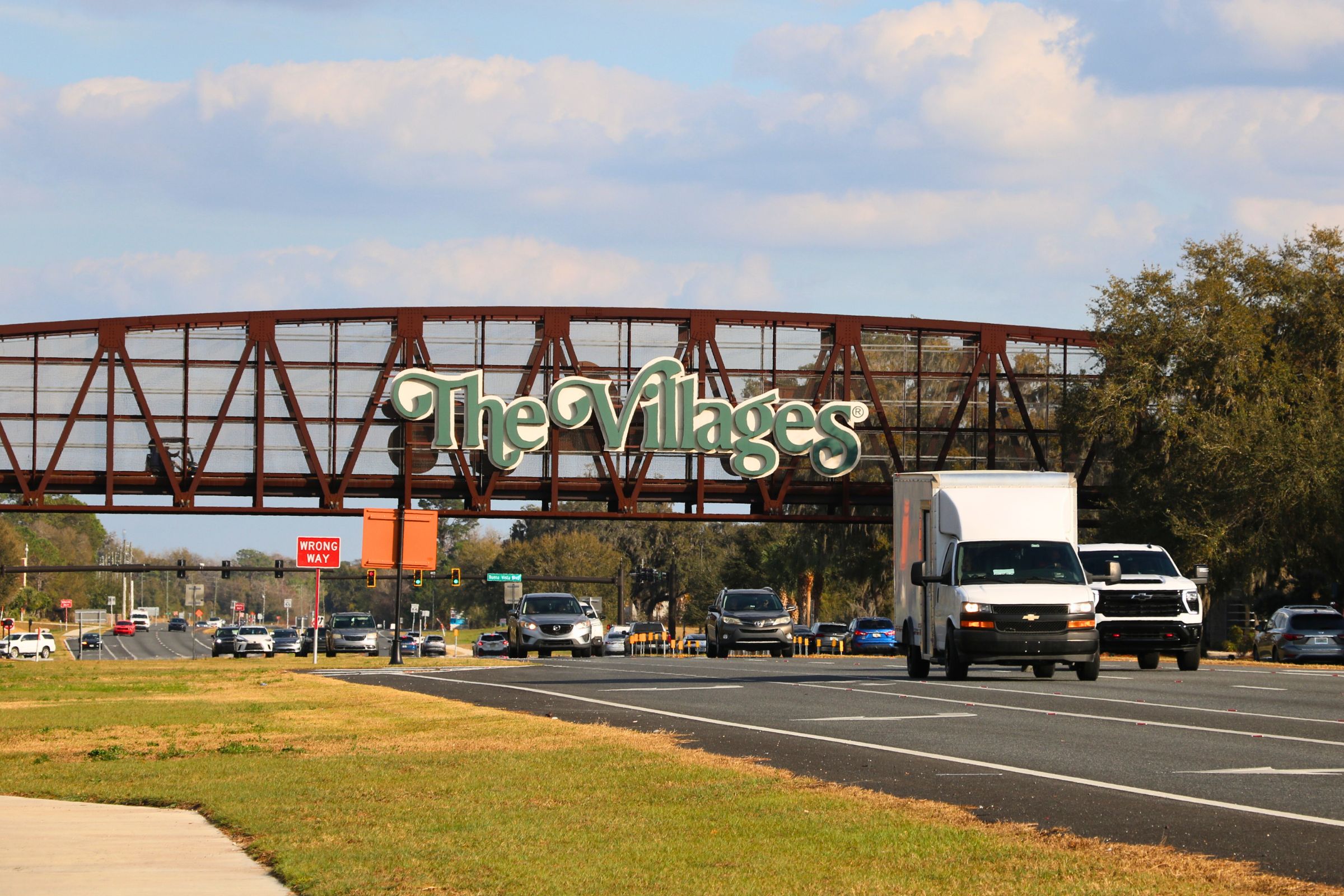 A brown, metal bridge for golf cart passing features large, green letters with white borders reading, "The Villages." A busy road passes underneath the bridge.