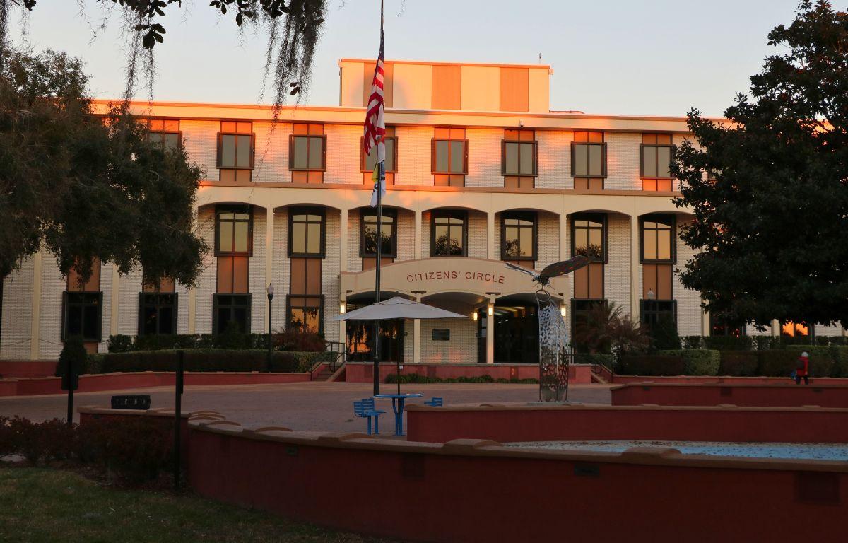 A large, brick building features stairs line with black metal railings and short, red walls. Over the entrance is a smaller overhang with red letters on the front reading, "citizen's circle." Picnic tables with umbrellas are around the area. A flagpole waves the American flag and the City of Ocala flag. A statue of a butterfly stands in the center. A small splash pad can be seen encircled by the red walls.