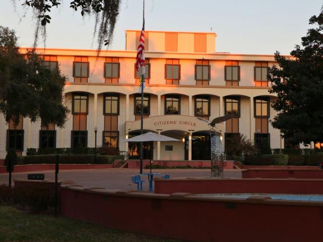 A large, brick building features stairs line with black metal railings and short, red walls. Over the entrance is a smaller overhang with red letters on the front reading, "citizen's circle." Picnic tables with umbrellas are around the area. A flagpole waves the American flag and the City of Ocala flag. A statue of a butterfly stands in the center. A small splash pad can be seen encircled by the red walls.