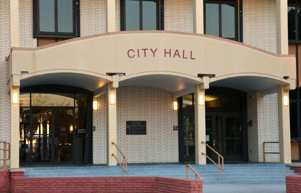 A large, brick building features an overhang over the entrance with red letters on the front reading, "City Hall." Stairs lead up into two double-door entrances to the building.