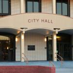 A large, brick building features an overhang over the entrance with red letters on the front reading, "City Hall." Stairs lead up into two double-door entrances to the building.