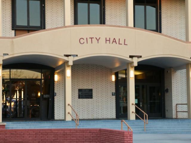 A large, brick building features an overhang over the entrance with red letters on the front reading, "City Hall." Stairs lead up into two double-door entrances to the building.