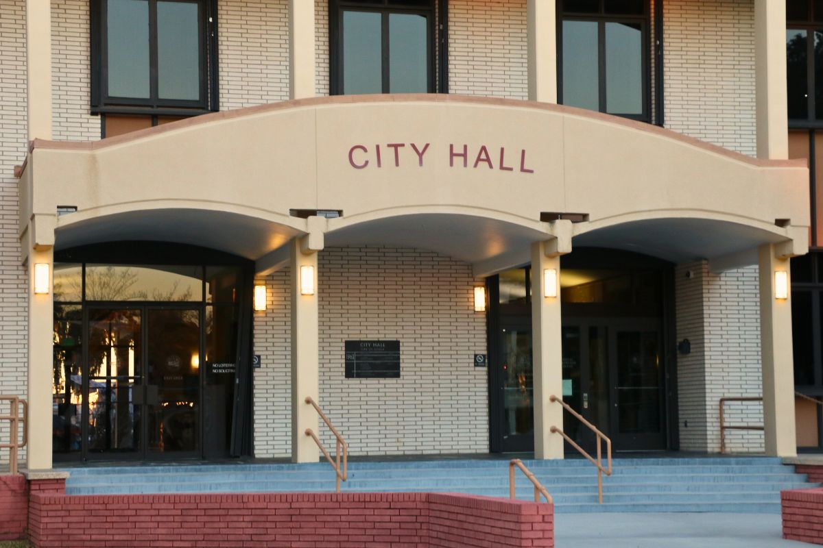 A large, brick building features an overhang over the entrance with red letters on the front reading, "City Hall." Stairs lead up into two double-door entrances to the building.