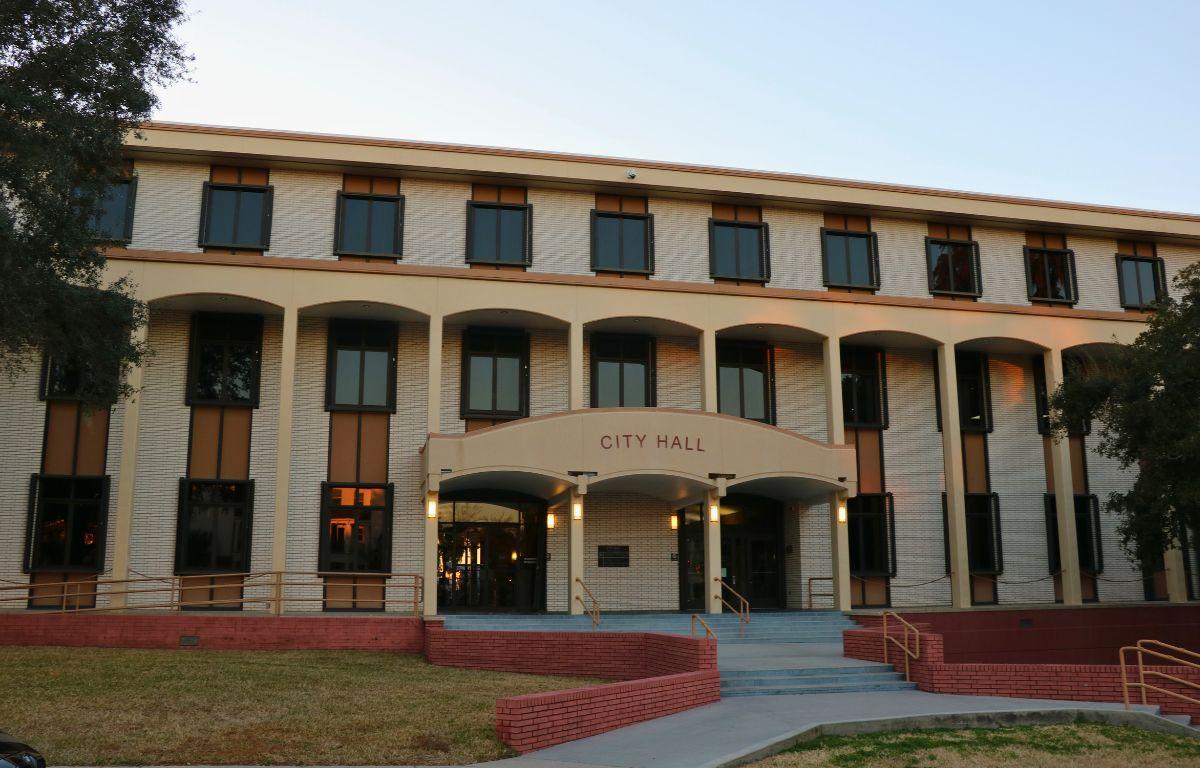 A large, brick building features stairs line with yellow metal railings and short, red brick walls. Over the entrance is a smaller overhang with red letters on the front reading, "city hall."