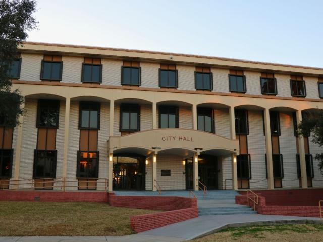 A large, brick building features stairs line with yellow metal railings and short, red brick walls. Over the entrance is a smaller overhang with red letters on the front reading, "city hall."