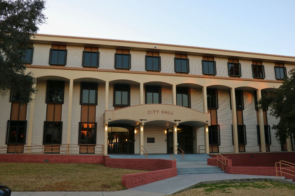 A large, brick building features stairs line with yellow metal railings and short, red brick walls. Over the entrance is a smaller overhang with red letters on the front reading, "city hall."