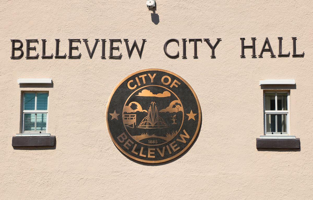A beige building features a large City of Belleview logo with small windows on either side of it. above the logo is letters reading, "Belleview City Hall." Above the letters is a small camera.