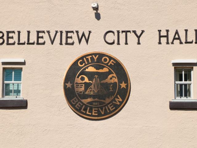 A beige building features a large City of Belleview logo with small windows on either side of it. above the logo is letters reading, "Belleview City Hall." Above the letters is a small camera.