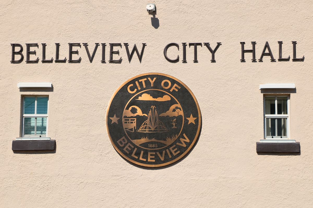 A beige building features a large City of Belleview logo with small windows on either side of it. above the logo is letters reading, "Belleview City Hall." Above the letters is a small camera.