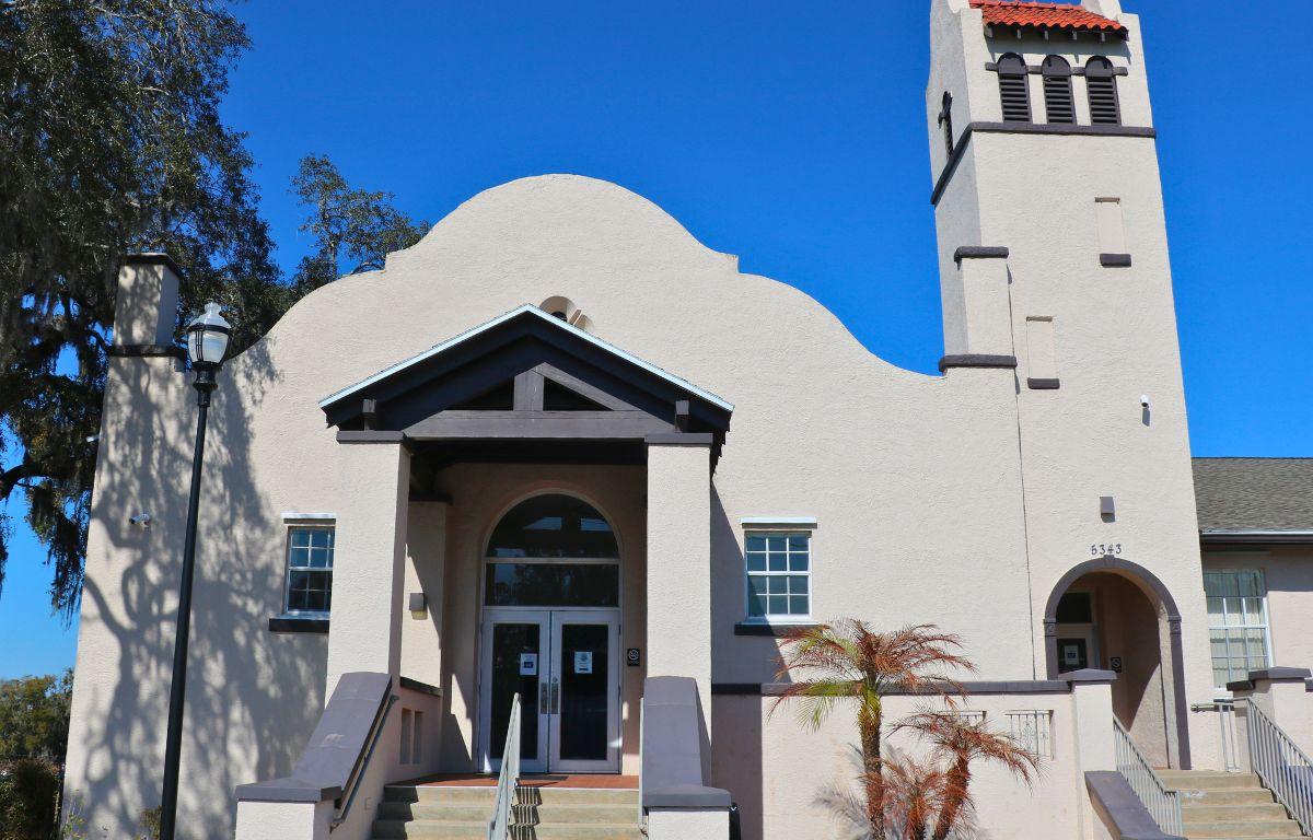 A white building with a decorative entrance features a tower to the right with another entrance at the bottom. Windows are on either side of the entrance with a large overhang between them over the doors. Stairs lead up to the doors on both entrances.