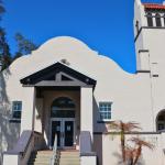 A white building with a decorative entrance features a tower to the right with another entrance at the bottom. Windows are on either side of the entrance with a large overhang between them over the doors. Stairs lead up to the doors on both entrances.