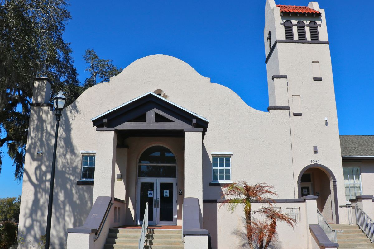 A white building with a decorative entrance features a tower to the right with another entrance at the bottom. Windows are on either side of the entrance with a large overhang between them over the doors. Stairs lead up to the doors on both entrances.