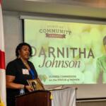 On the left an American flag, the flag of the State of Florida, a woman behind a podium with a microphone, a large screen to the woman's right with writing and an image.