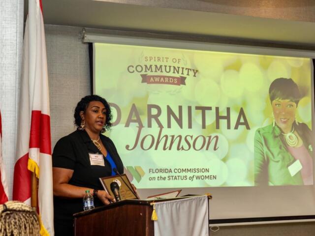 On the left an American flag, the flag of the State of Florida, a woman behind a podium with a microphone, a large screen to the woman's right with writing and an image.