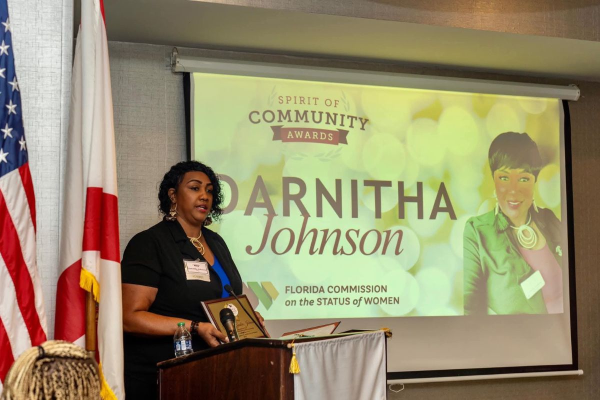 On the left an American flag, the flag of the State of Florida, a woman behind a podium with a microphone, a large screen to the woman's right with writing and an image.