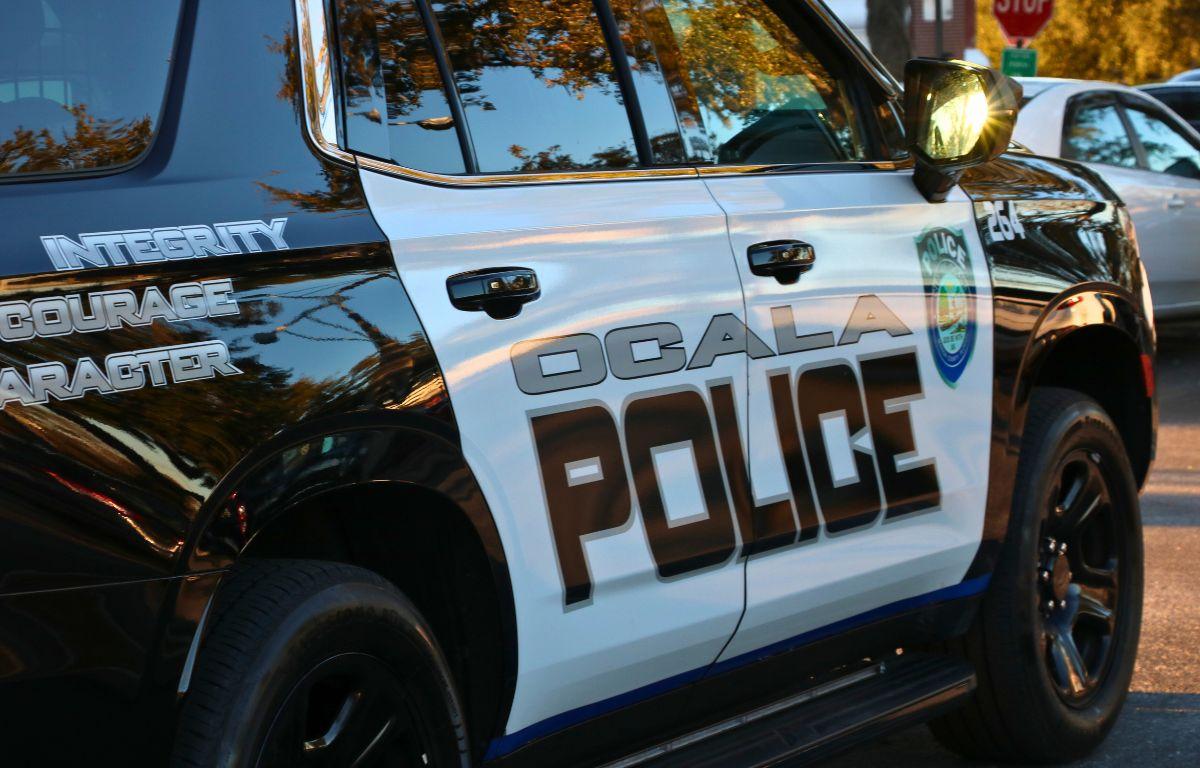 A black and white police car features text reading, "Ocala Police" and "Integrity. Courage. Character." Toward the front of the car is a City of Ocala police logo. In the background, more cars can be seen in a parking lot. The sun reflects off the mirror.