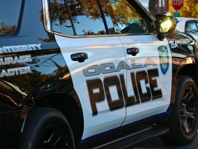 A black and white police car features text reading, "Ocala Police" and "Integrity. Courage. Character." Toward the front of the car is a City of Ocala police logo. In the background, more cars can be seen in a parking lot. The sun reflects off the mirror.