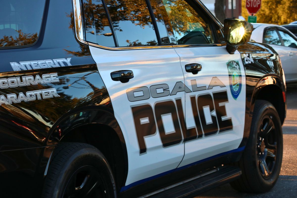 A black and white police car features text reading, "Ocala Police" and "Integrity. Courage. Character." Toward the front of the car is a City of Ocala police logo. In the background, more cars can be seen in a parking lot. The sun reflects off the mirror.
