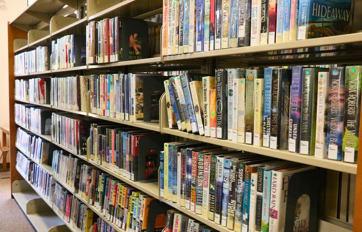 Books are lined up on a wooden seven-tired bookshelf in a brightly lit library.