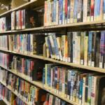 Books are lined up on a wooden seven-tired bookshelf in a brightly lit library.