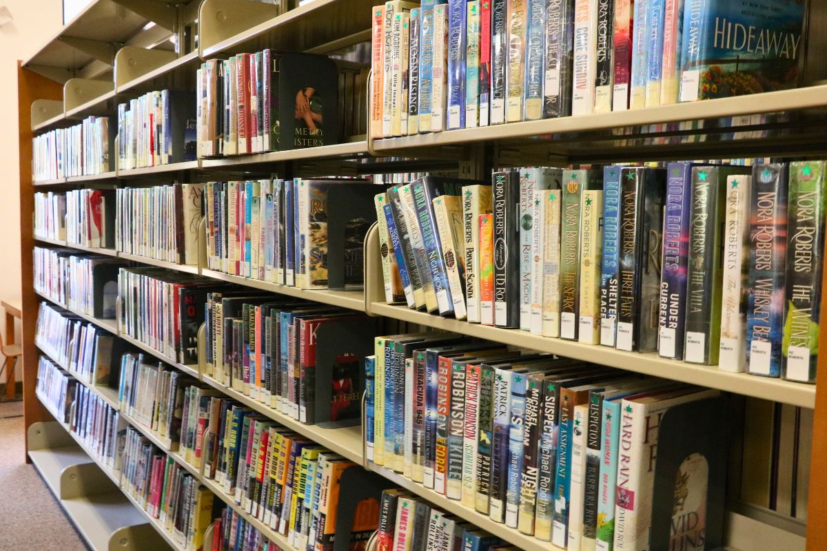 Books are lined up on a wooden seven-tired bookshelf in a brightly lit library.