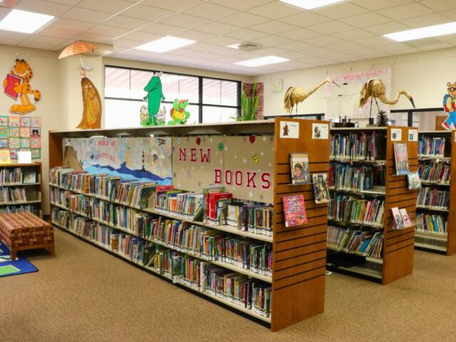Five shelves are lined with books in a children's reading area. Stickers on the wall and characters on the shelves display fictional characters found in children's books and cartoons.
