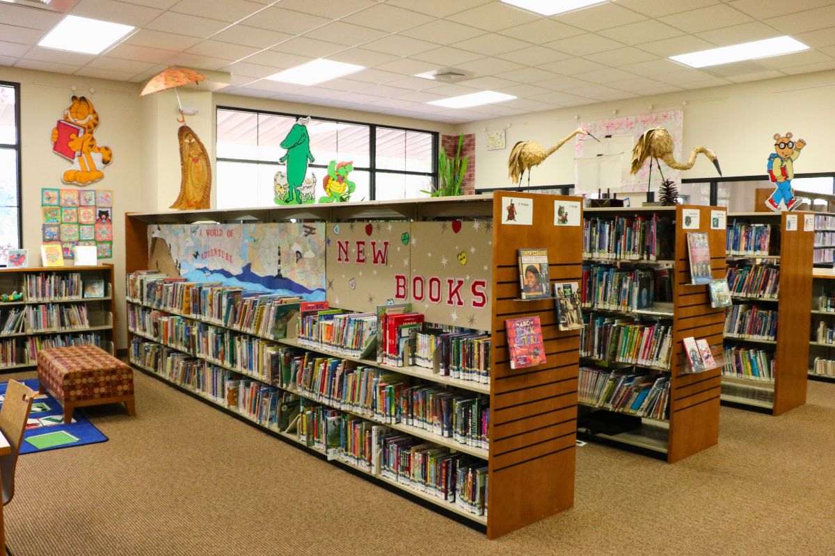 Five shelves are lined with books in a children's reading area. Stickers on the wall and characters on the shelves display fictional characters found in children's books and cartoons.