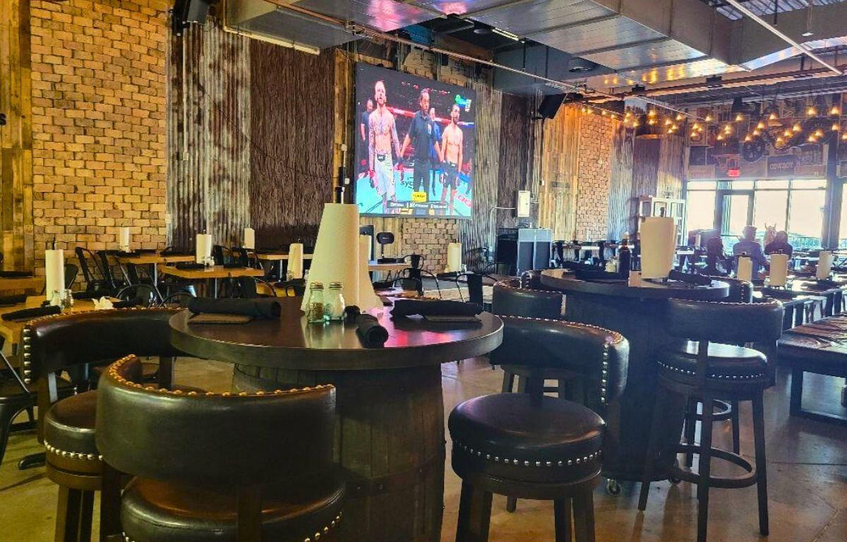 A restaurant features barrel-shaped tables with bar stools around them. A large RV screen hangs in the background showing a boxing match. More wooden tables are scattered around the restaurant.