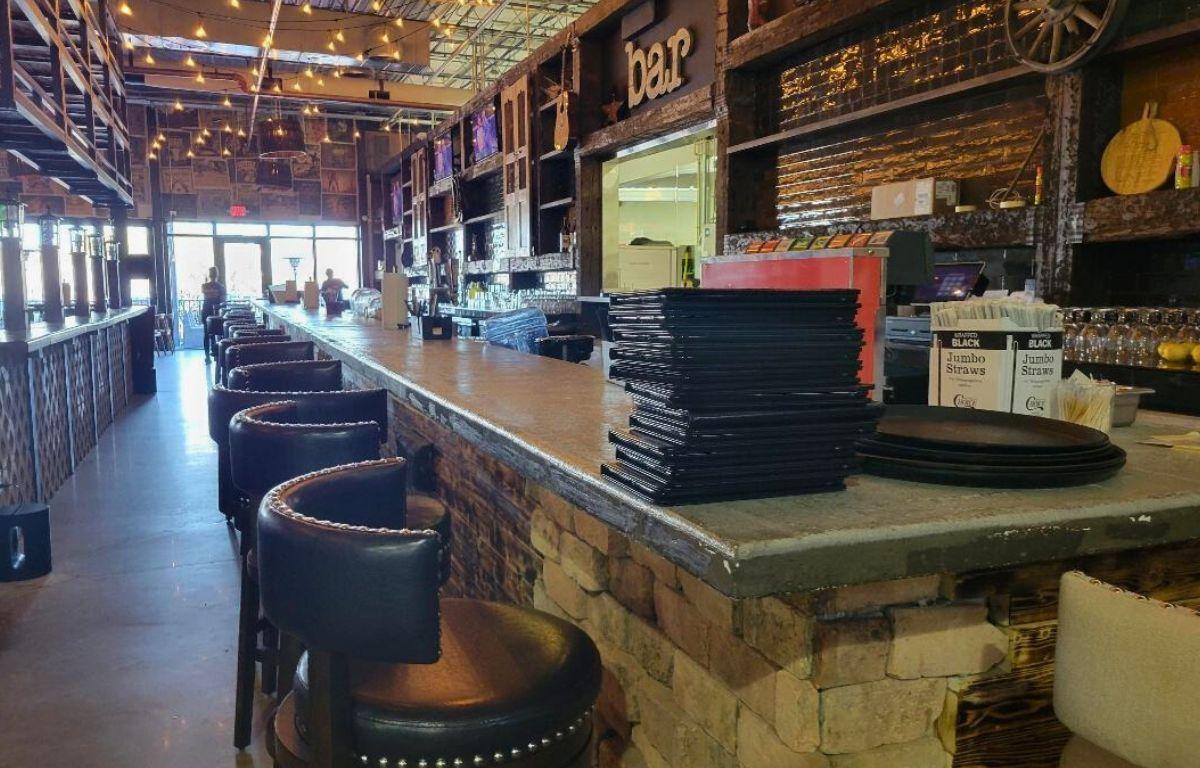 A lengthy, rustic-looking bar stands toward the back of restaurant, featuring seating along the countertops and a stack of menus toward the corner.