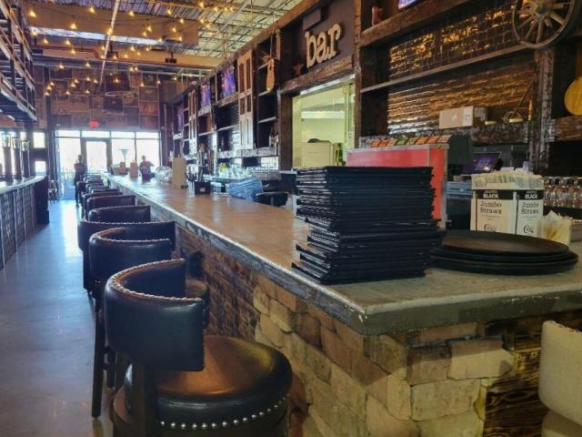 A lengthy, rustic-looking bar stands toward the back of restaurant, featuring seating along the countertops and a stack of menus toward the corner.