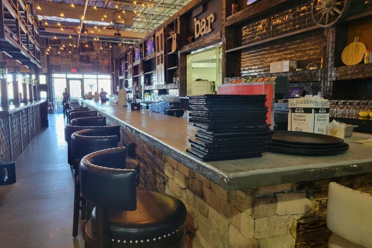 A lengthy, rustic-looking bar stands toward the back of restaurant, featuring seating along the countertops and a stack of menus toward the corner.