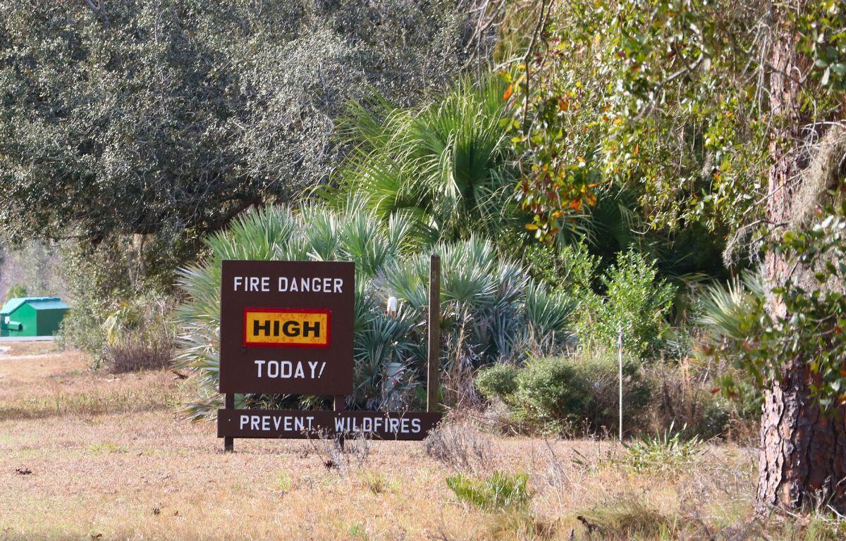 A wood sign features white text reading, "Fire Danger Today! Prevent Wildfires." In the center of the sign is a yellow box with a red border with black text reading, "High." The sign is placed in a forest.