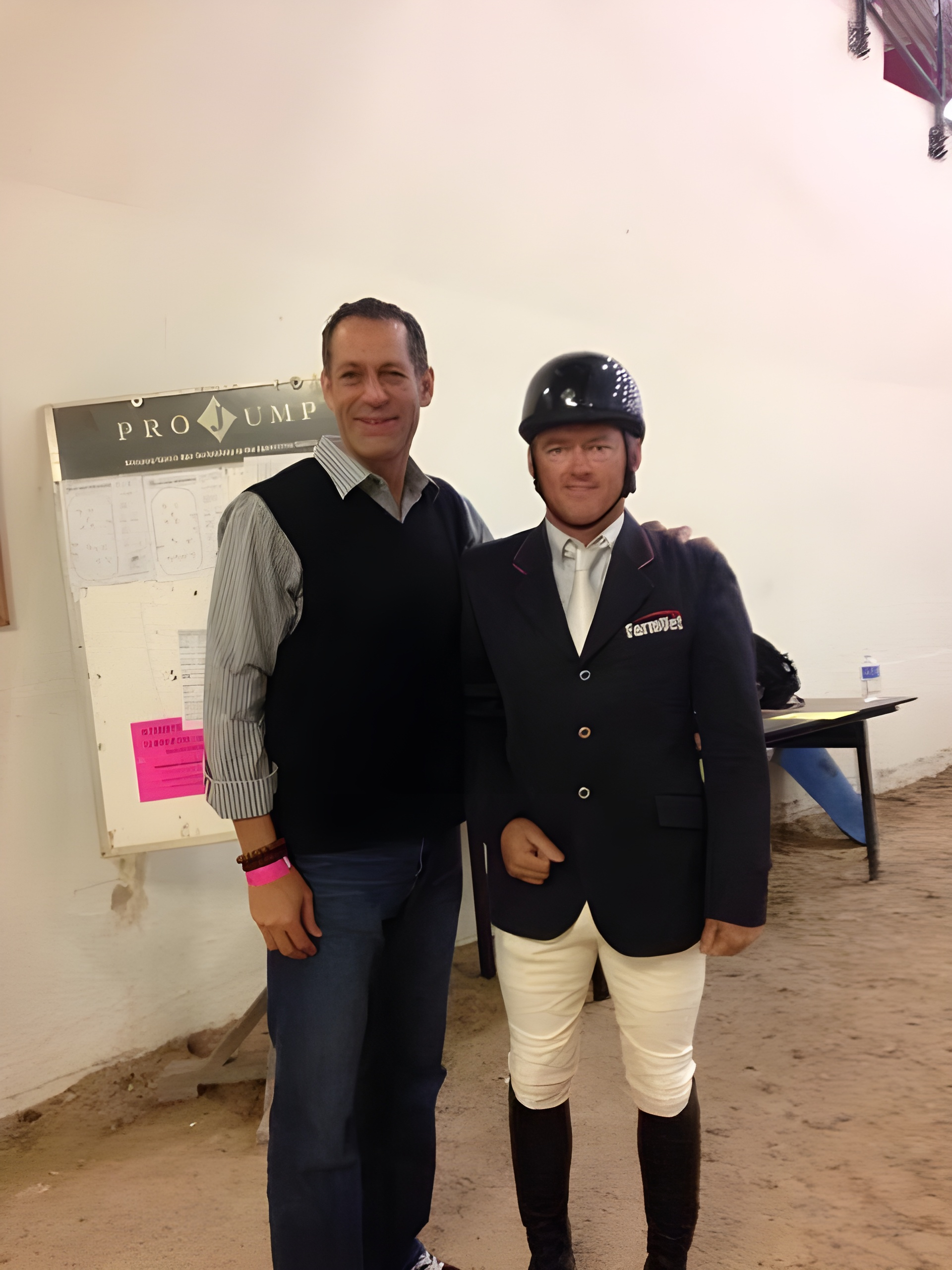 Two men standing together, the one on the left wearing a vest, one on the right wearing a riding jacket. An easel in the back holding a placard in front of a wall. 