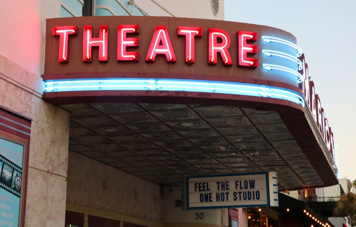 A theatre features a large, light up marquee that reads, "Theatre." The marquee is on an overhang. Hanging from the bottom is a white and blue sign that reads, "Feel the flow one hot studio.