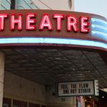 A theatre features a large, light up marquee that reads, "Theatre." The marquee is on an overhang. Hanging from the bottom is a white and blue sign that reads, "Feel the flow one hot studio.