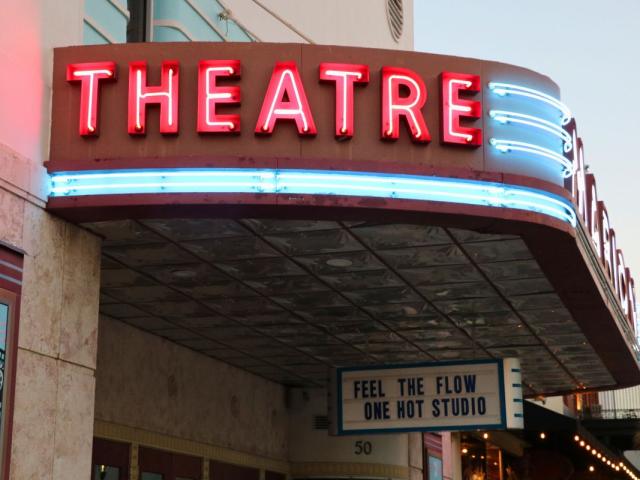 A theatre features a large, light up marquee that reads, "Theatre." The marquee is on an overhang. Hanging from the bottom is a white and blue sign that reads, "Feel the flow one hot studio.