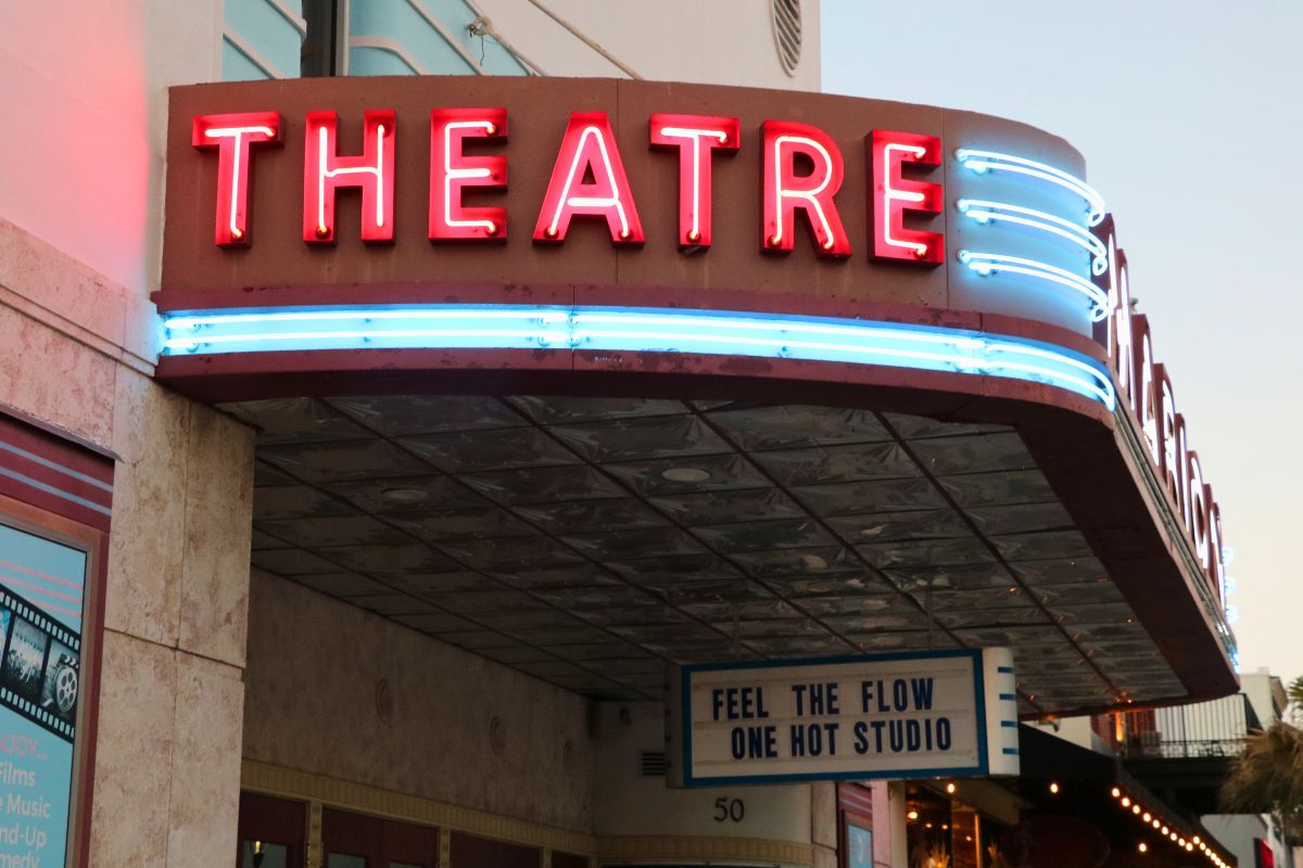 A theatre features a large, light up marquee that reads, "Theatre." The marquee is on an overhang. Hanging from the bottom is a white and blue sign that reads, "Feel the flow one hot studio.