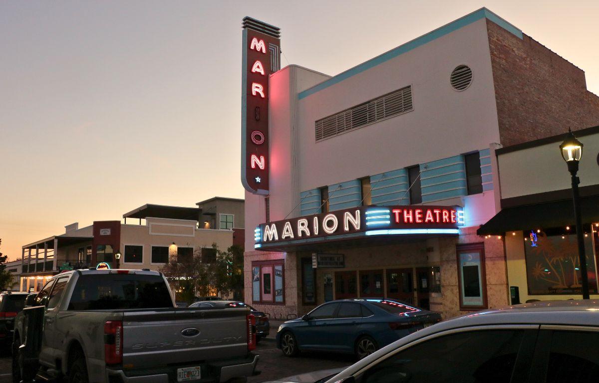 A large theatre sits on a street corner with cars parked along the street in front of and across the street from theatre. The theatre has large, light up signs reading, "Marion Theatre."