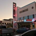 A large theatre sits on a street corner with cars parked along the street in front of and across the street from theatre. The theatre has large, light up signs reading, "Marion Theatre."
