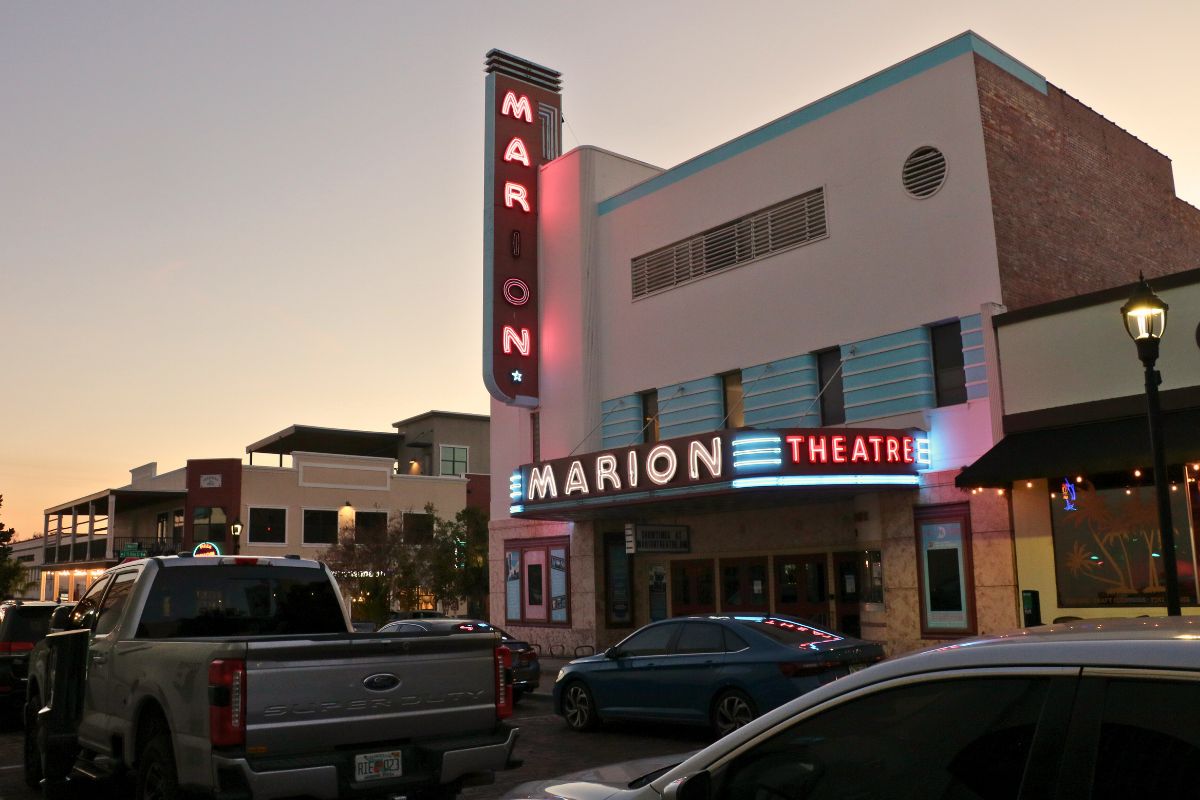 A large theatre sits on a street corner with cars parked along the street in front of and across the street from theatre. The theatre has large, light up signs reading, "Marion Theatre."