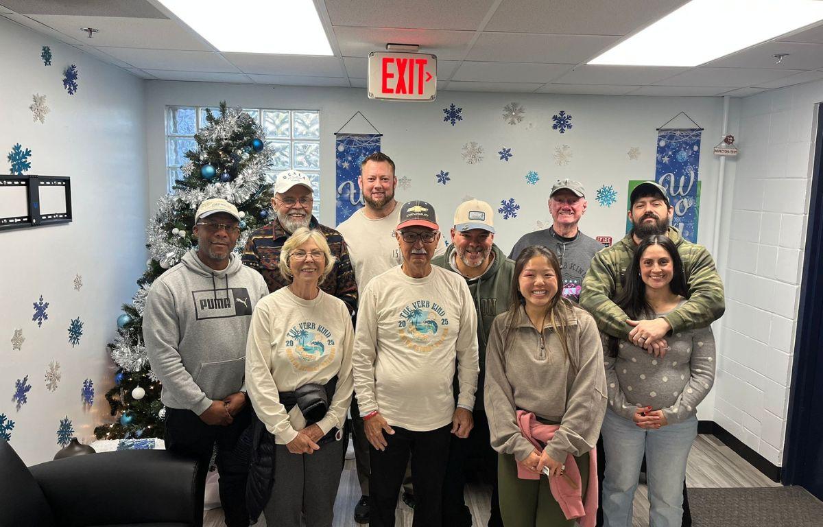 Christmas tree in the background, blue and white on the wall in the background, with two rows of people standing in front of and next to one another.