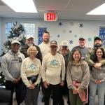 Christmas tree in the background, blue and white on the wall in the background, with two rows of people standing in front of and next to one another.