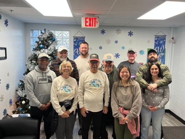 Christmas tree in the background, blue and white on the wall in the background, with two rows of people standing in front of and next to one another.