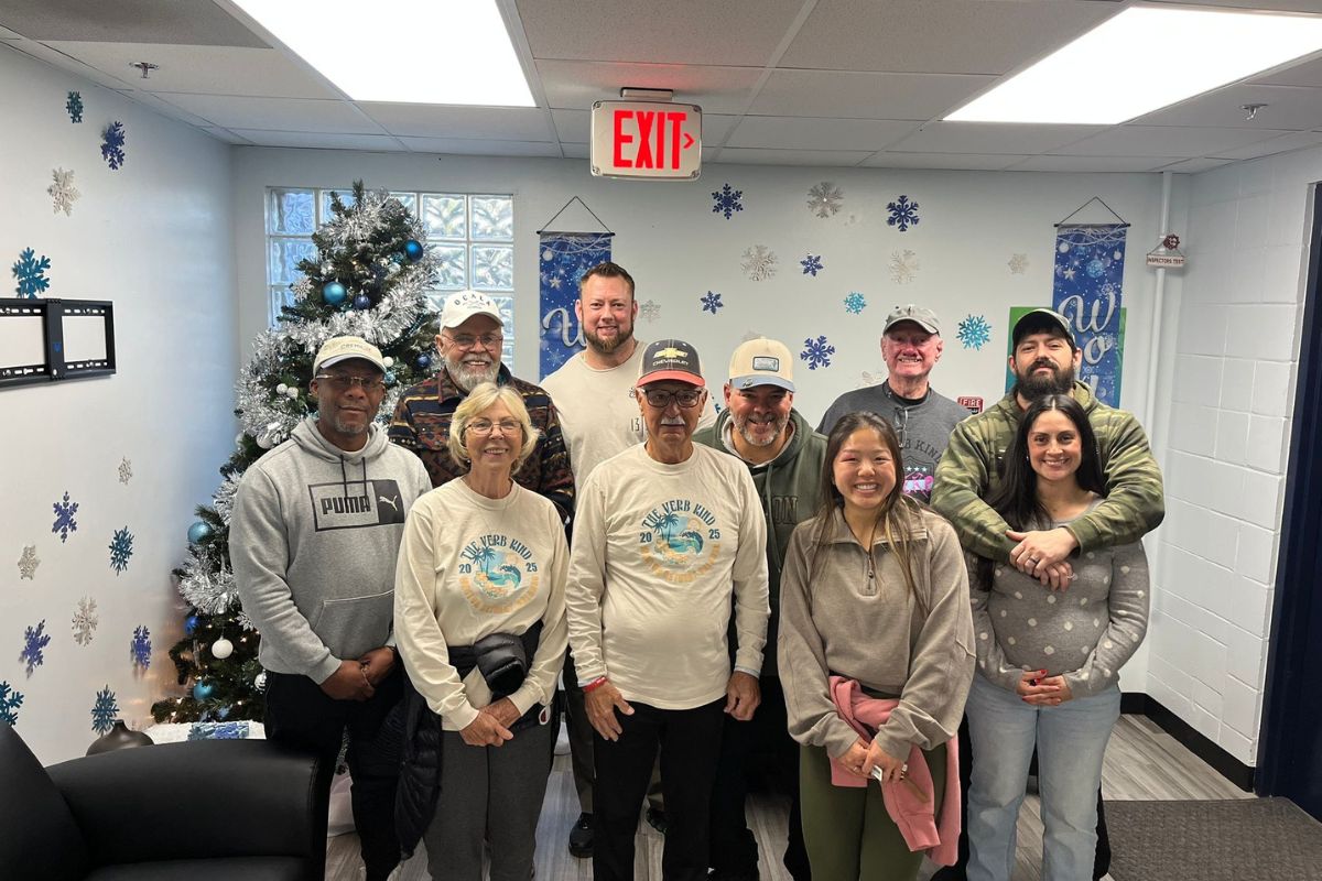 Christmas tree in the background, blue and white on the wall in the background, with two rows of people standing in front of and next to one another.