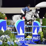 A horsewoman on in a blue blazer riding a gray horse clear a jumping effort, with MARS Standard, with a canopy in the background.