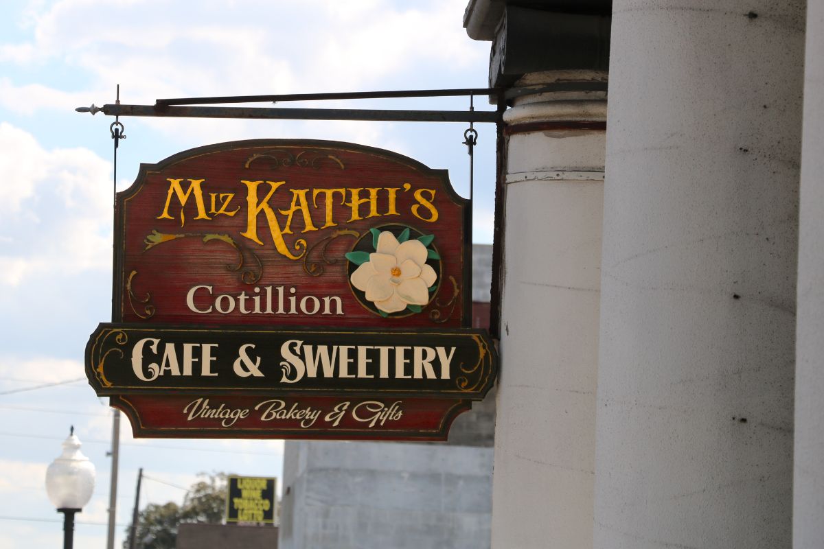 A sign hangs from a pillar with a vintage look reading, "Miz Kathi's Cotillion Cafe & Sweetery. Vintage Bakery & Gifts."