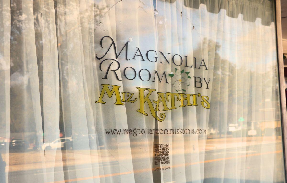 A window with white curtains behind it has a sticker in the front reading, "Magnolia Room by Miz Kathi's. www.magnoliaroom.mizkathys.com." In the reflection is a street with full parking lot on the other side.
